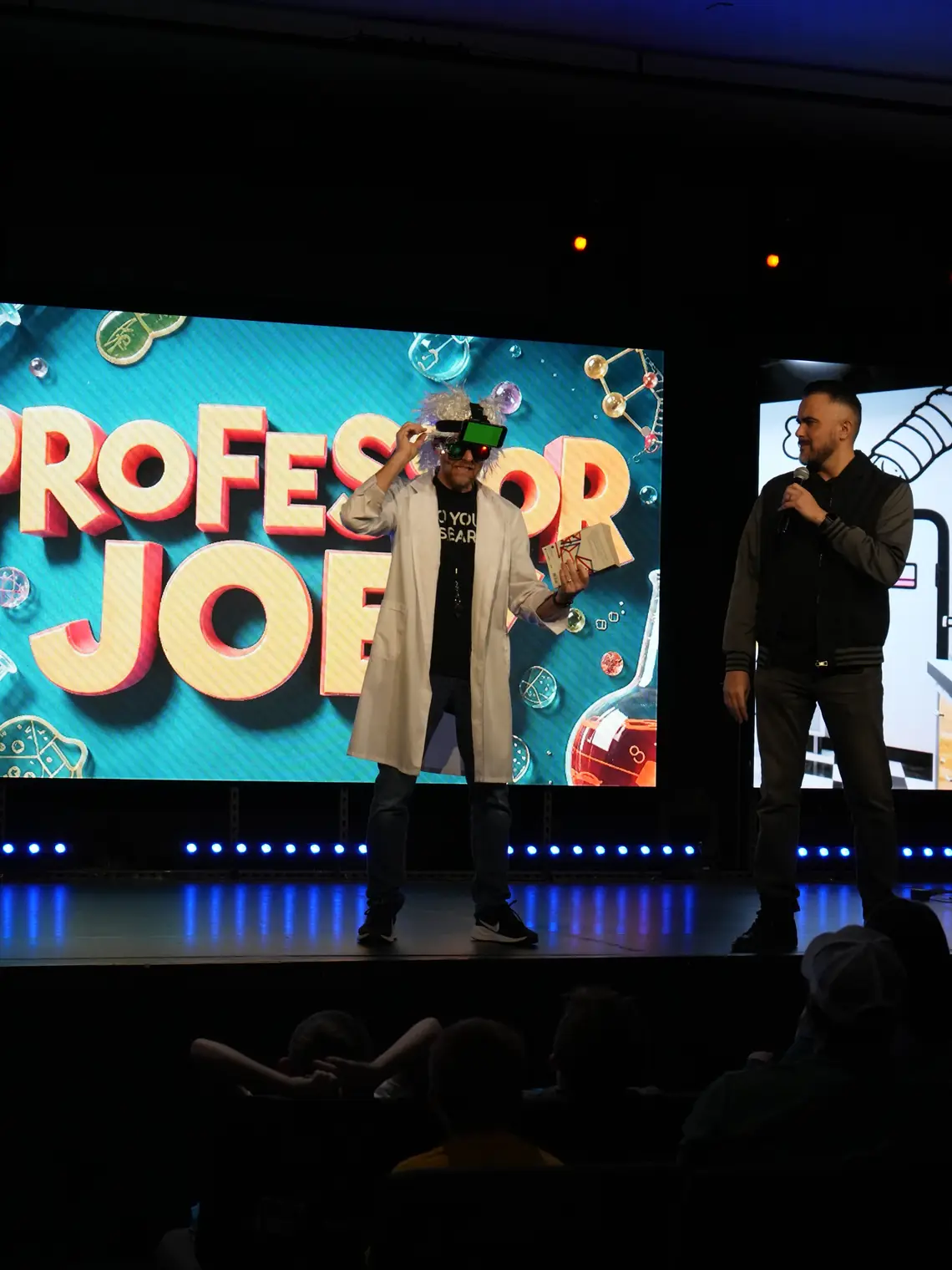 Professor Joey and the host on stage in front of a huge Professor Joey screen, with kids watching from the floor.