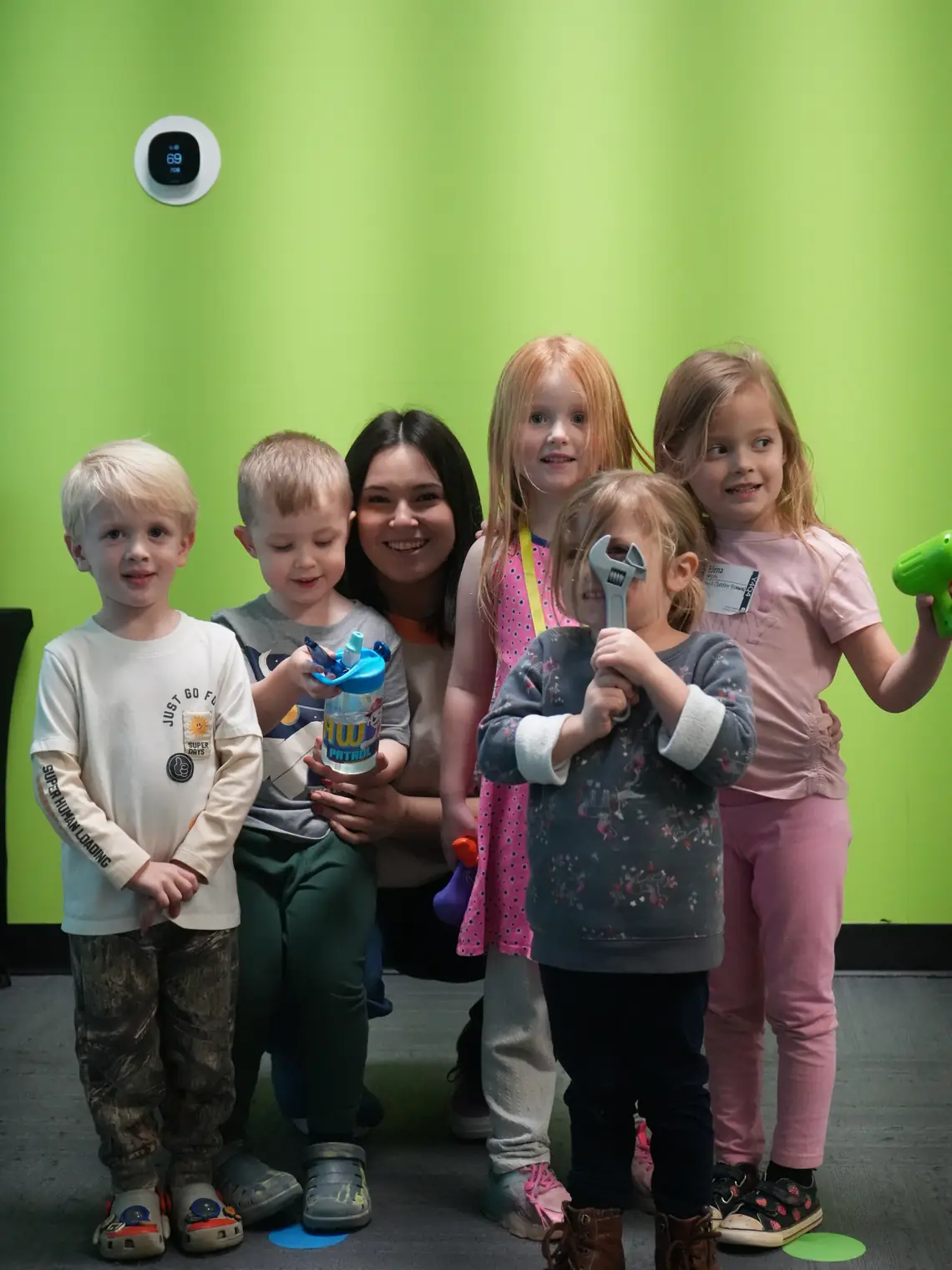 A leader crouched with five preschoolers against a bright green wall, all looking at the camera.